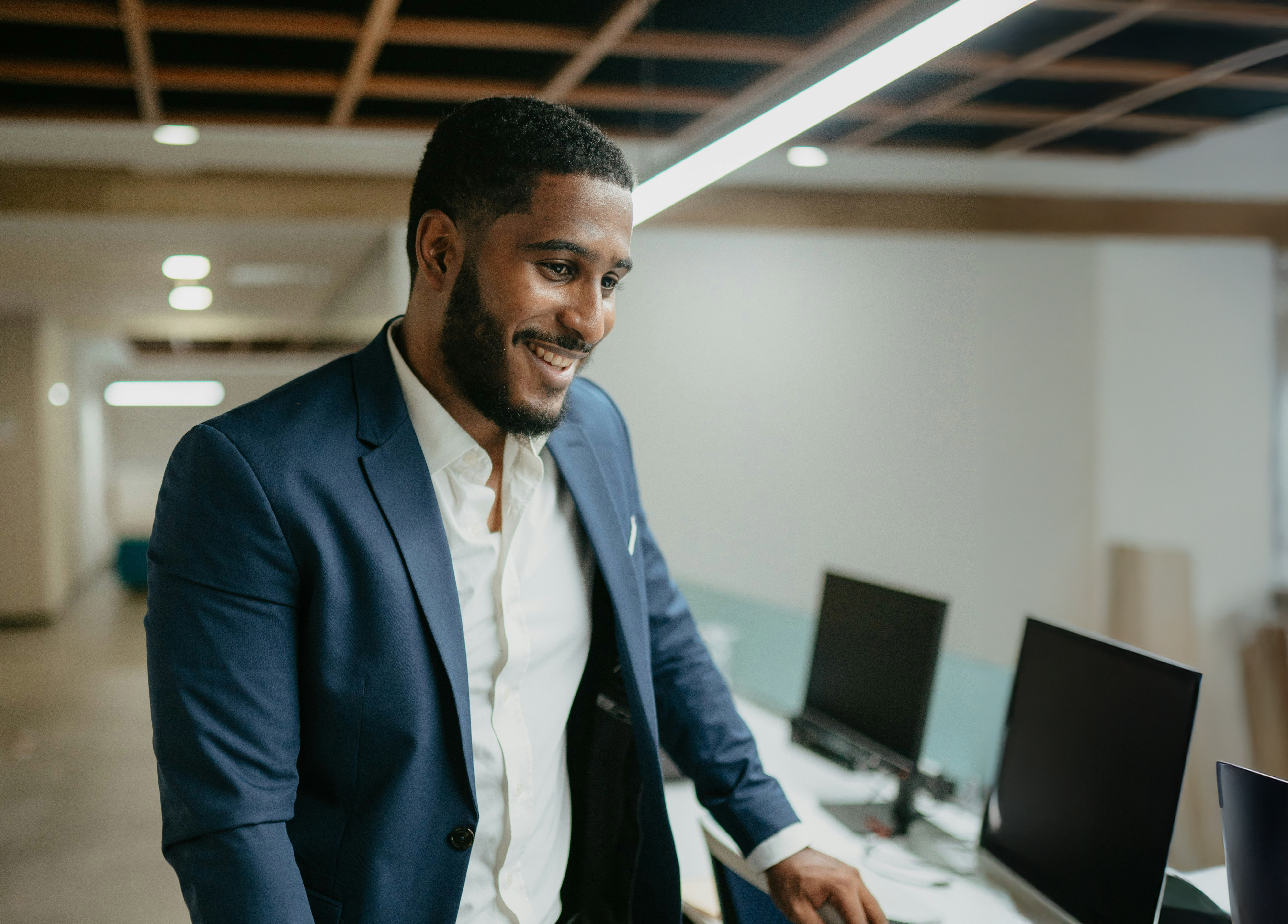 Smiling man in navy suit and white shirt standing near desks with computers in office.