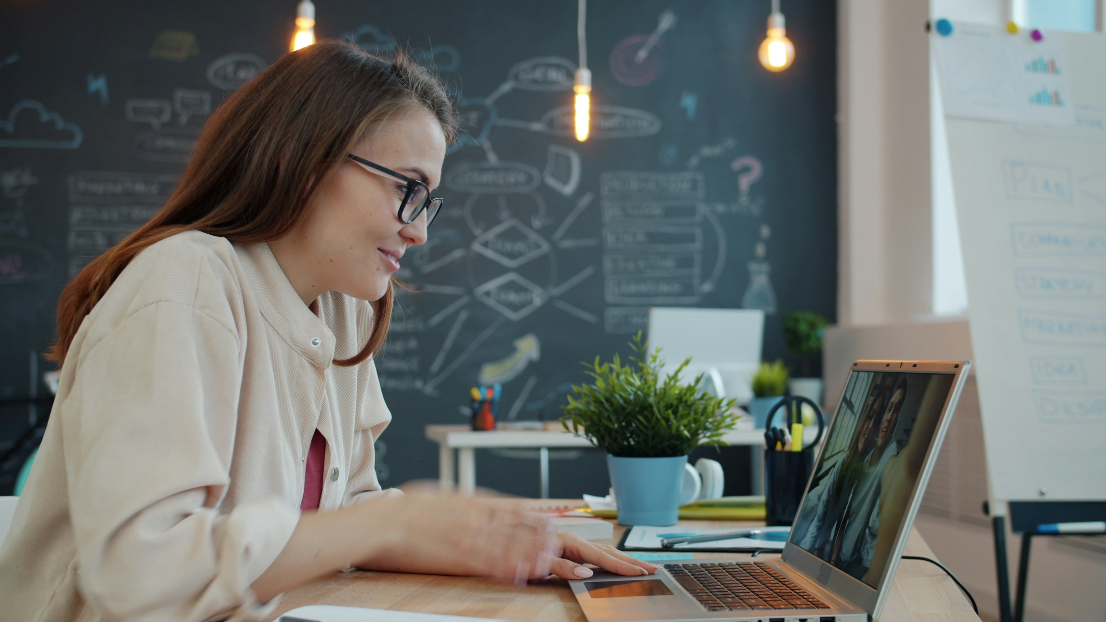 Woman with glasses smiling while video chatting on laptop at a bright office desk.