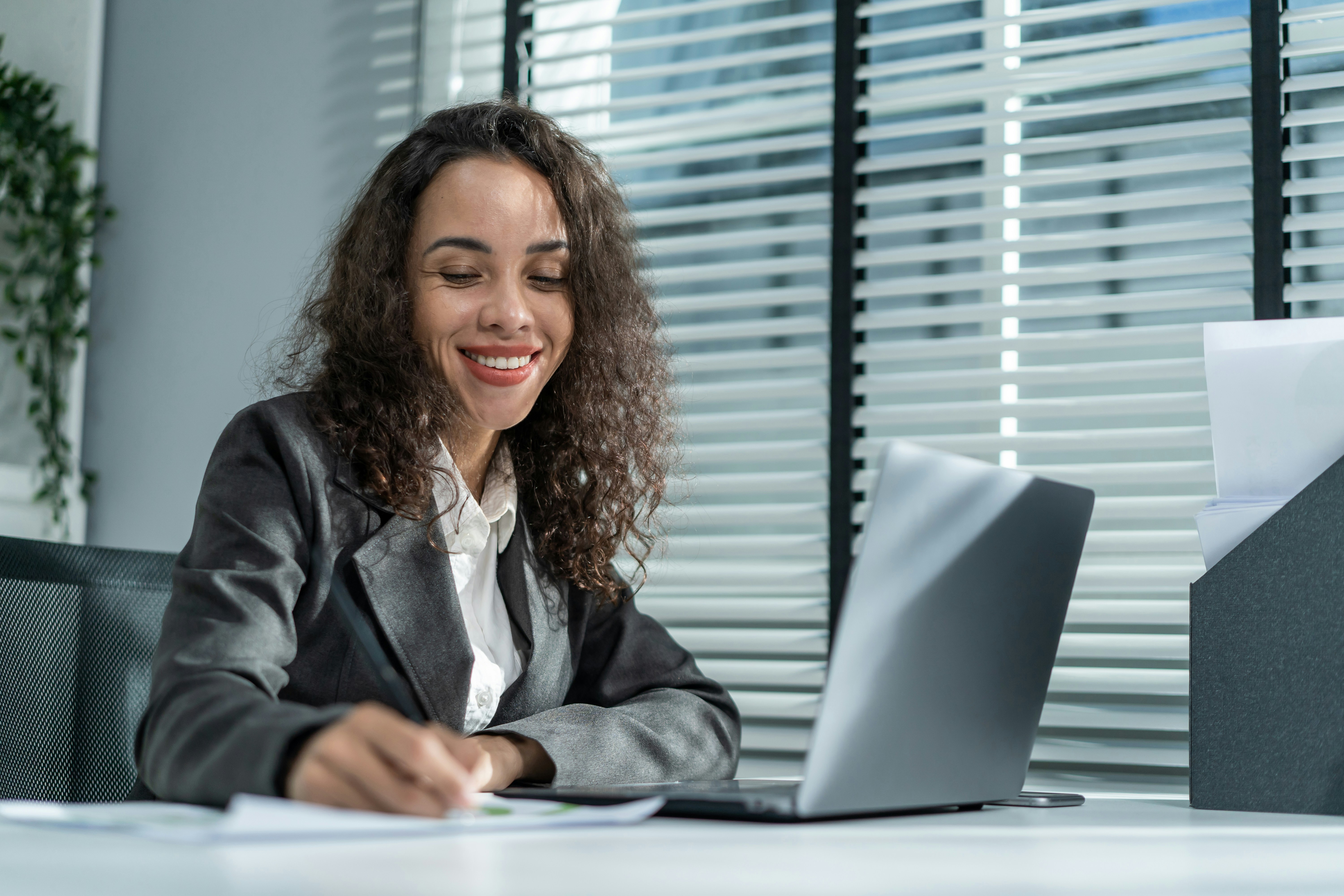 Smiling businesswoman writing on paper at desk with laptop in bright office with blinds.