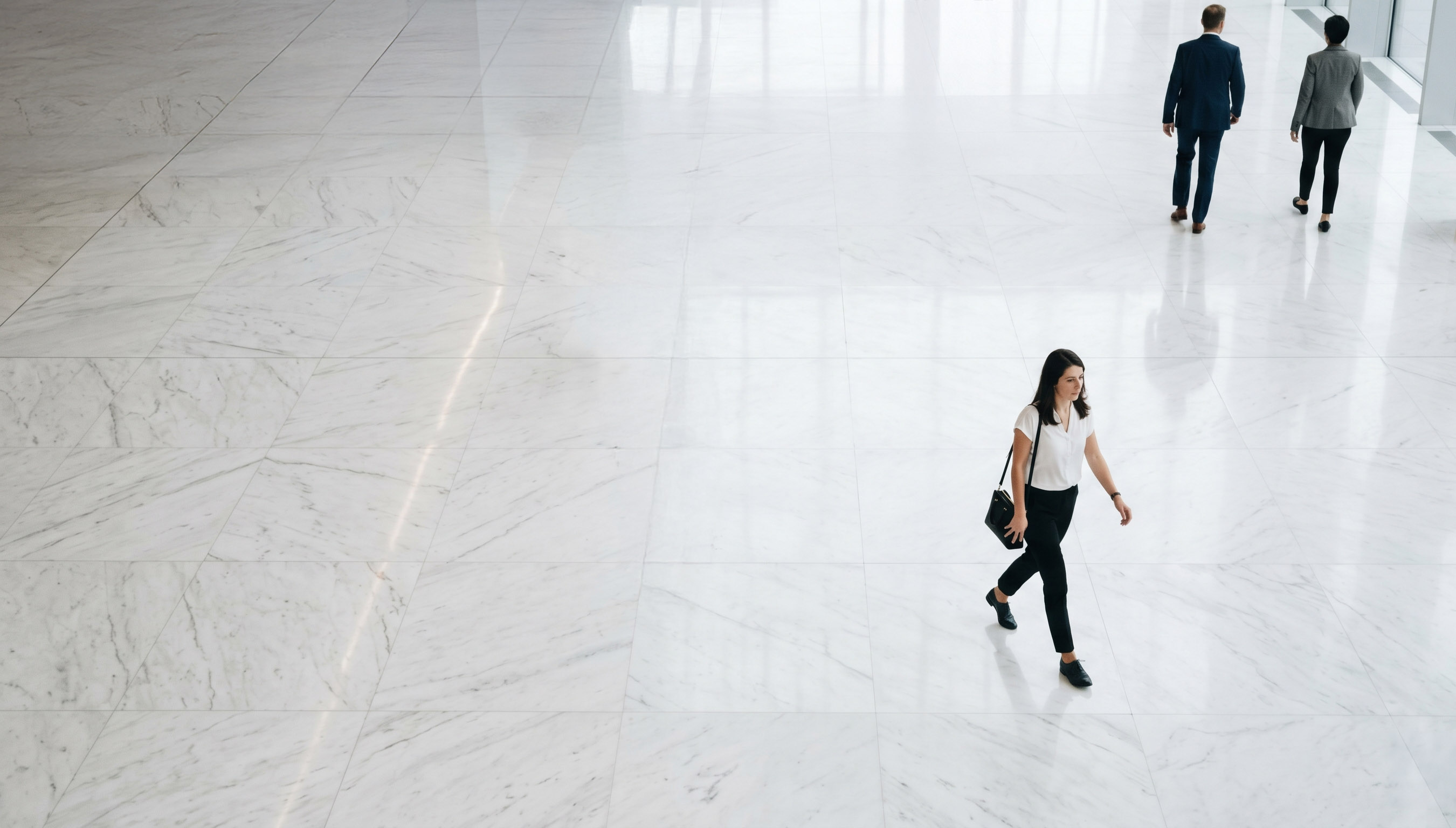Three professionally dressed people walking in a spacious white marble indoor area with large windows.