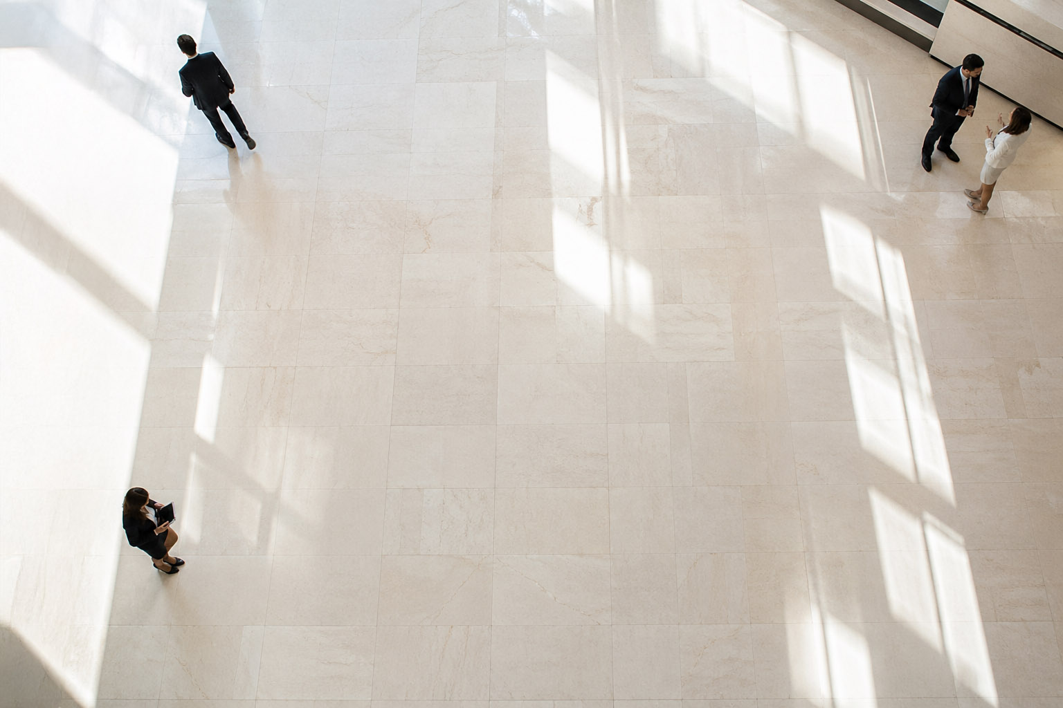 Aerial view of four business people standing apart on a large tiled floor with window shadows.