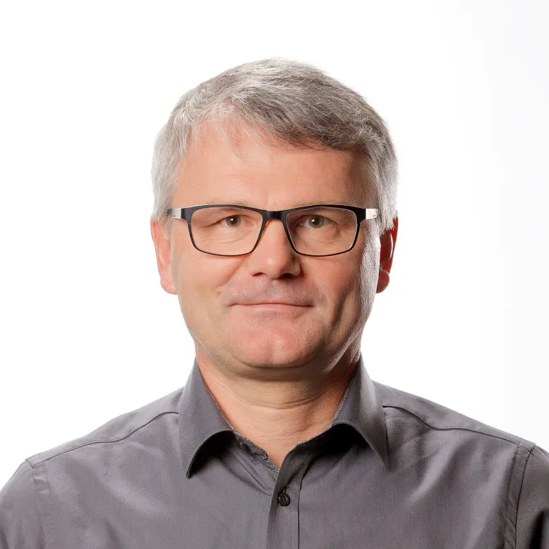 Homme d'âge mûr aux cheveux gris et portant des lunettes, vêtu d'une chemise grise sur fond blanc.