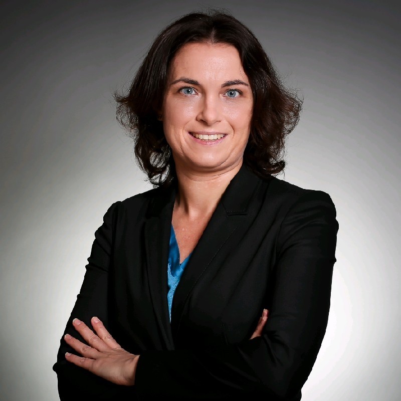 Professional woman with brown hair smiling, arms crossed, in black blazer and blue top on grey background.