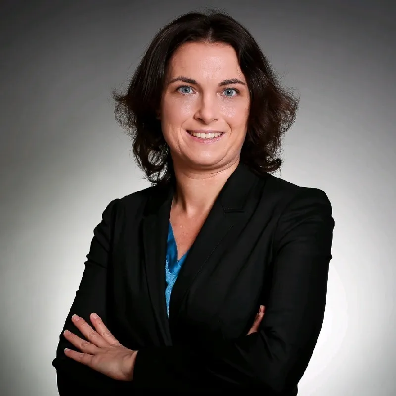 Femme professionnelle aux cheveux bruns, souriante, les bras croisés, vêtue d'un blazer noir et d'un haut bleu sur fond gris.