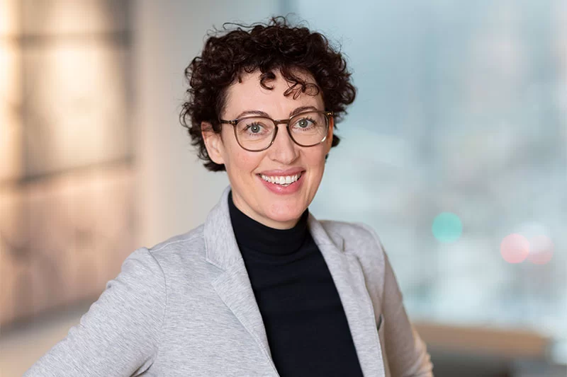 Smiling woman with short curly hair wearing glasses, black top, and light grey blazer in an office setting.