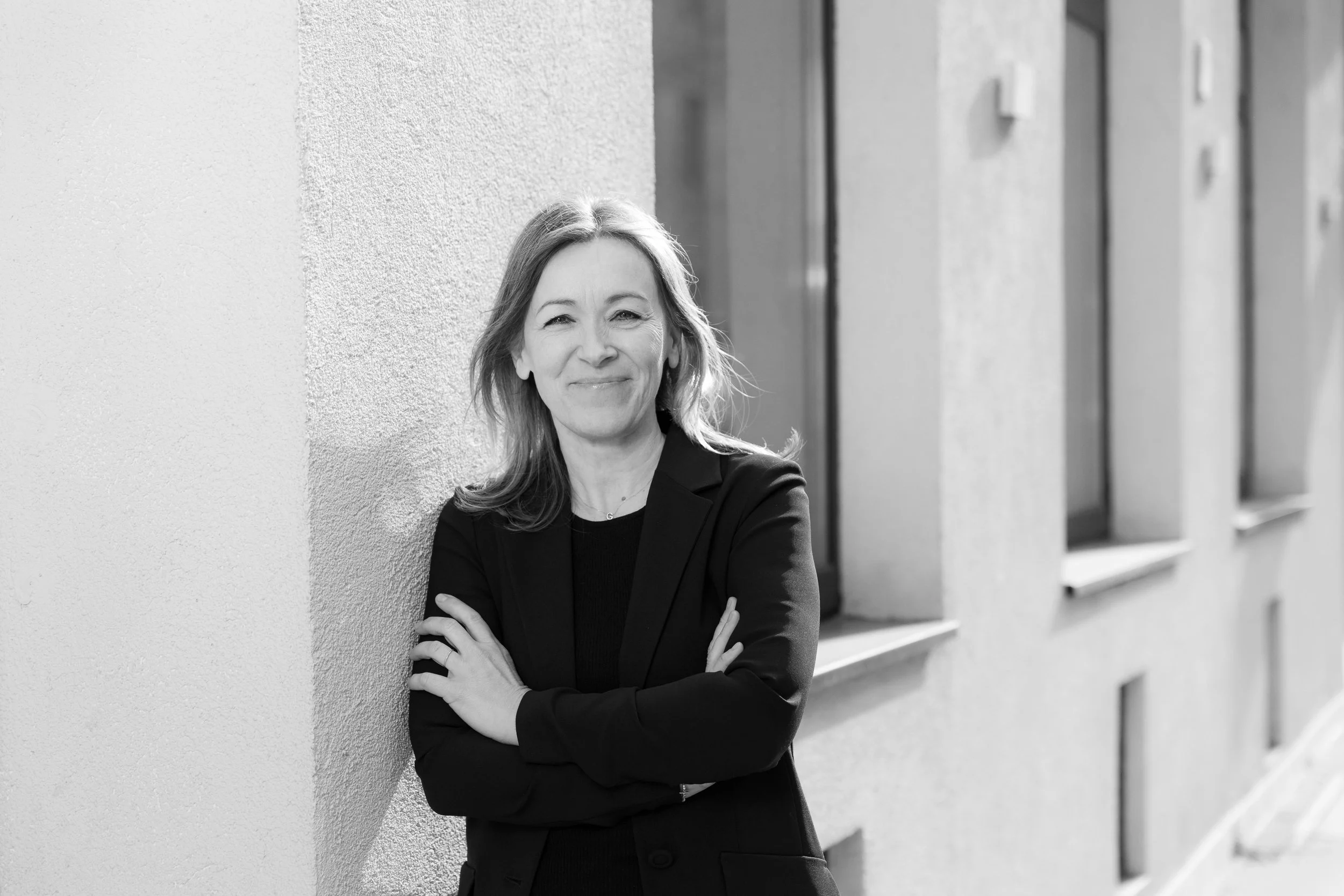 Smiling woman in black jacket poses with crossed arms against a bright building wall.