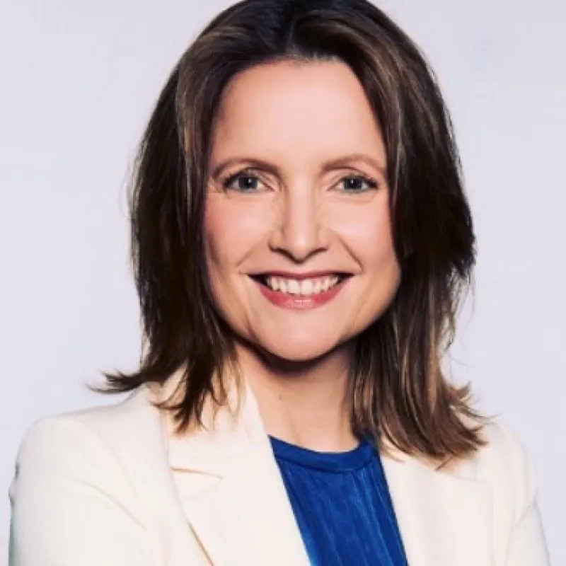 Femme souriante aux cheveux bruns mi-longs, portant un blazer blanc et un haut bleu.