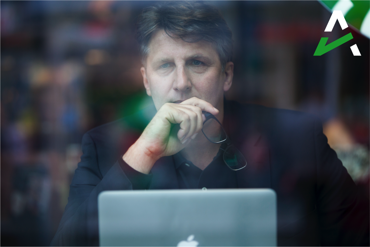 Man in black jacket holding glasses, looking thoughtful behind a window with a laptop in front of him.