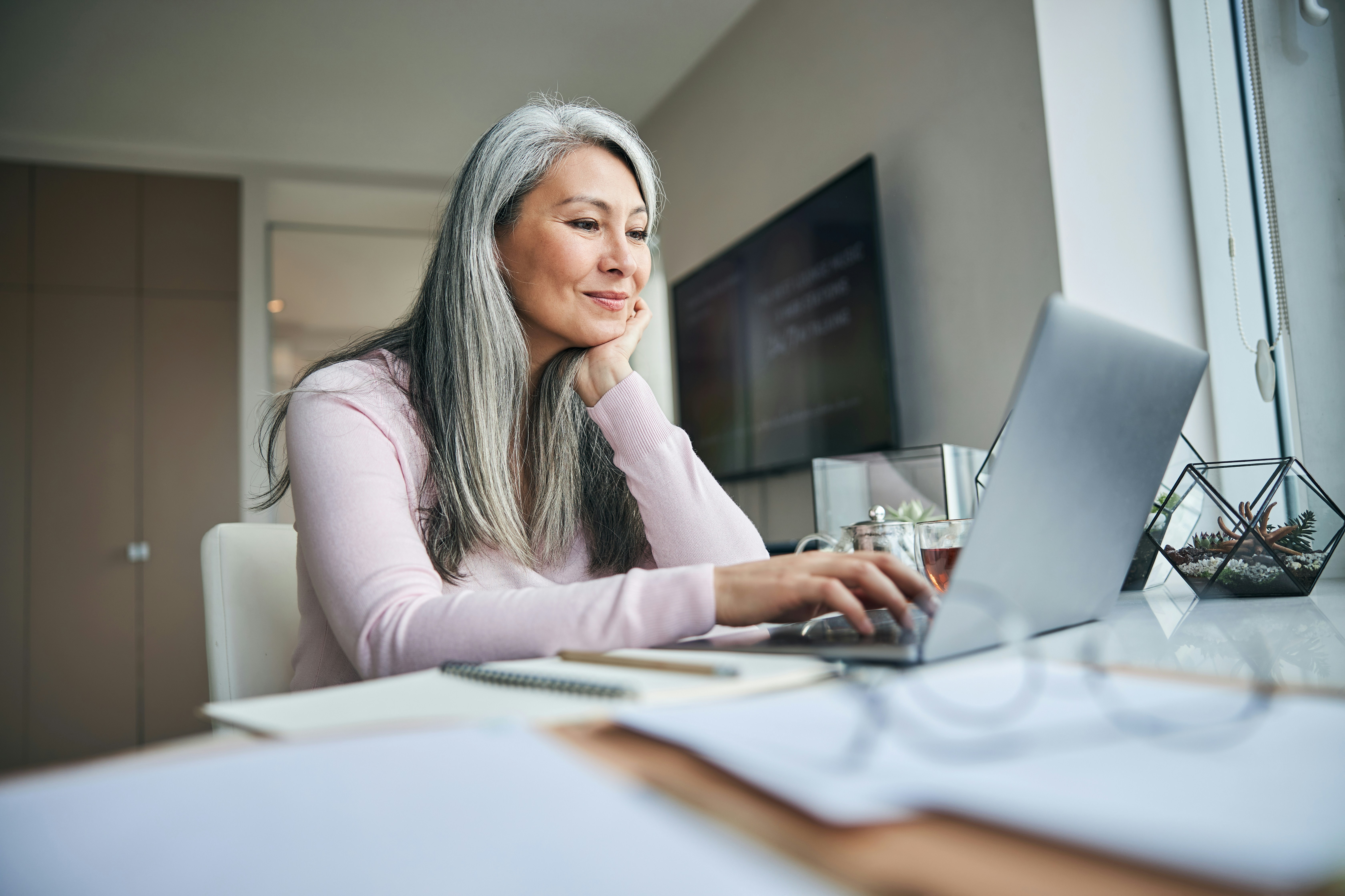 Mature woman with grey hair sitting at desk typing on laptop in bright modern room near window.