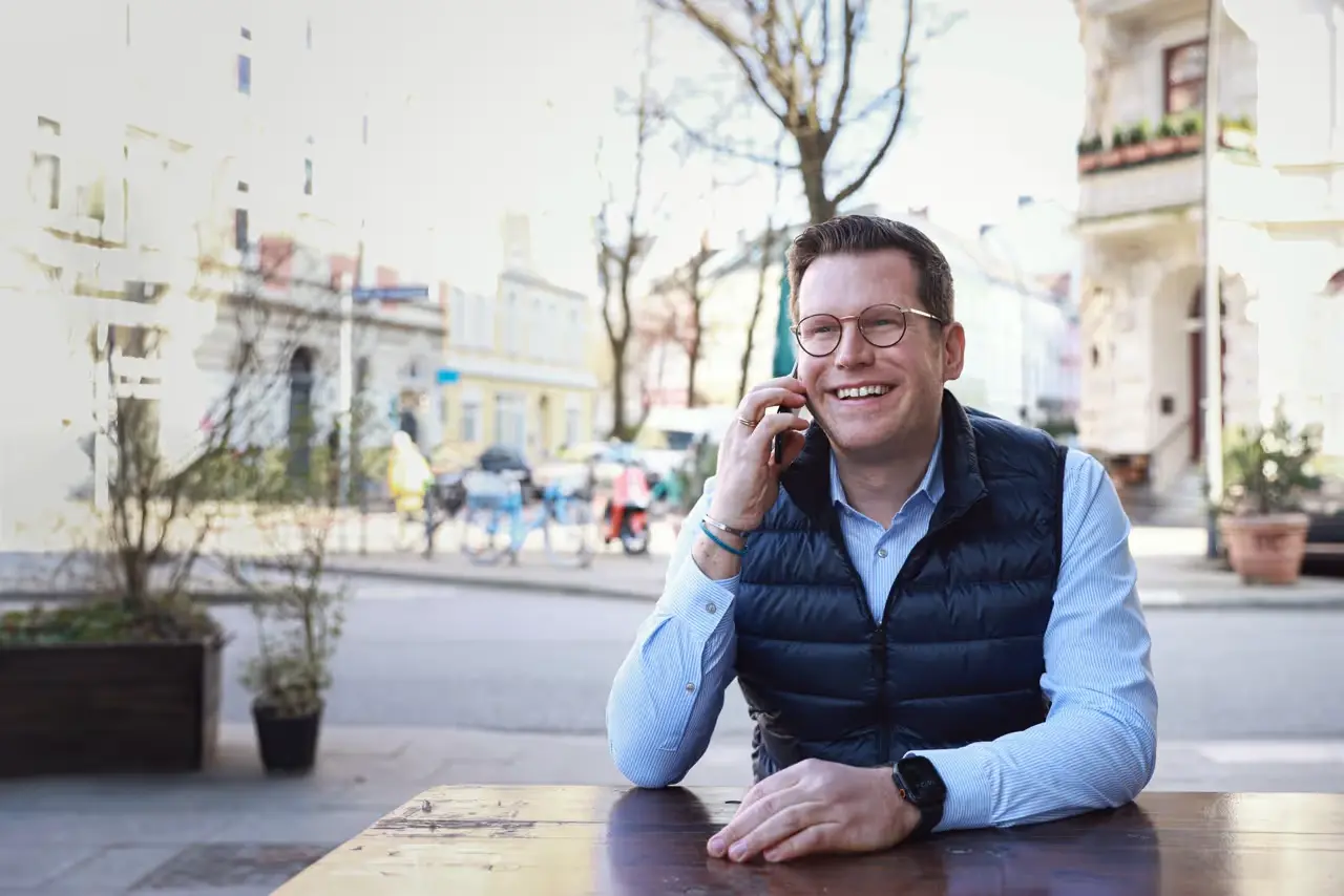 Un homme souriant, portant des lunettes et un gilet noir, parle au téléphone à une table en terrasse dans la rue.