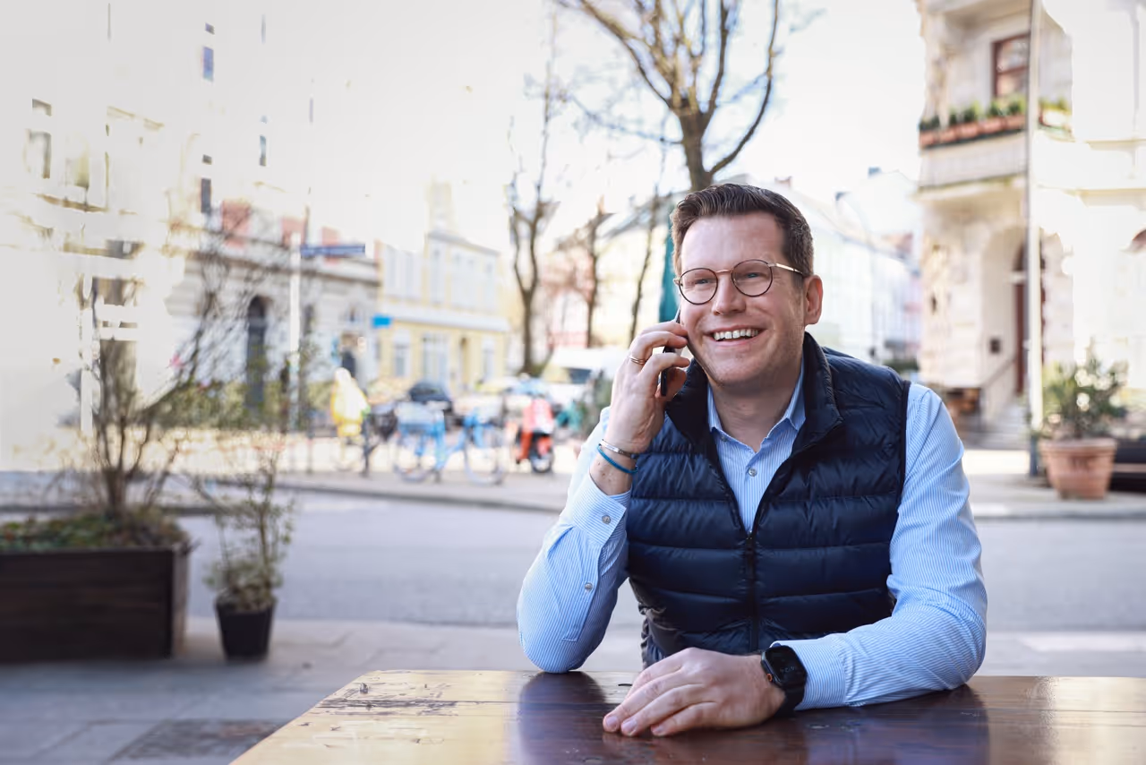 Smiling man wearing glasses and a black vest talking on a phone at an outdoor table on a street.