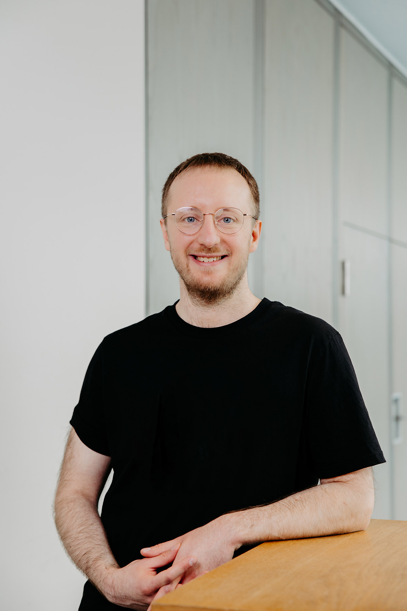 Lächelnder Mann mit Brille und Bart in schwarzem T-Shirt, der sich drinnen auf eine Holzfläche stützt