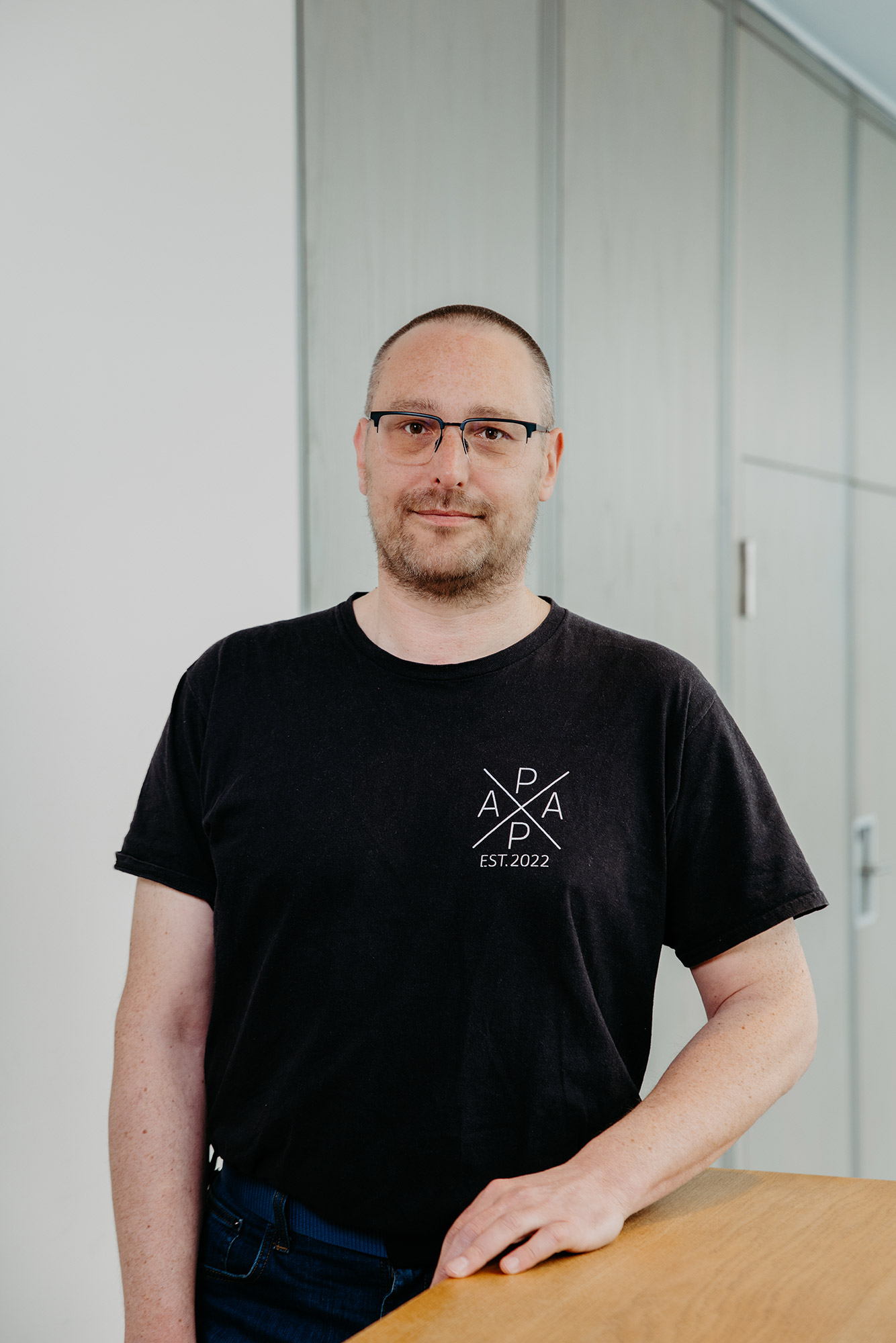 Mann mit Brille im schwarzen T-Shirt steht an einem Holztisch vor hellem Hintergrund