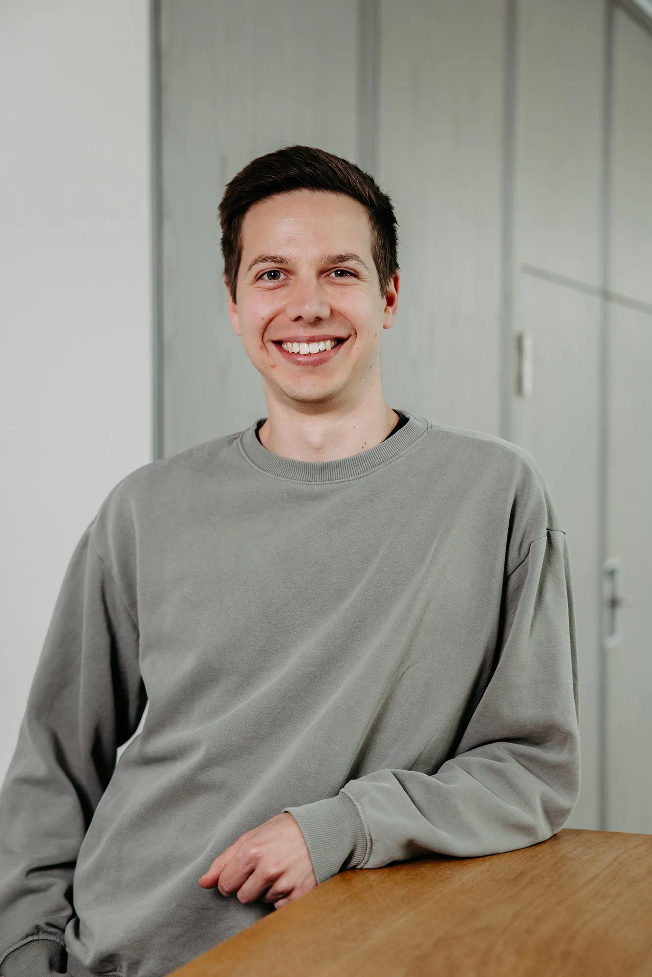 Lächelnder junger Mann im grauen Sweatshirt, an einen Holztisch gelehnt vor hellgrauem Hintergrund