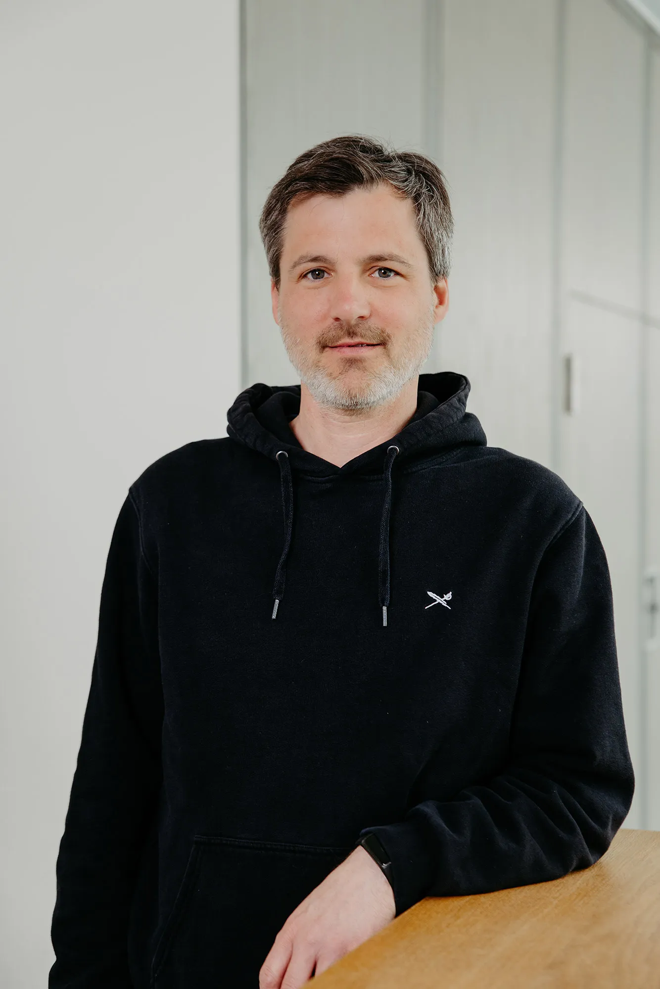 Mann mit kurzen dunklen Haaren und grauem Bart in schwarzem Hoodie und Smartwatch, an einen Holztisch gelehnt