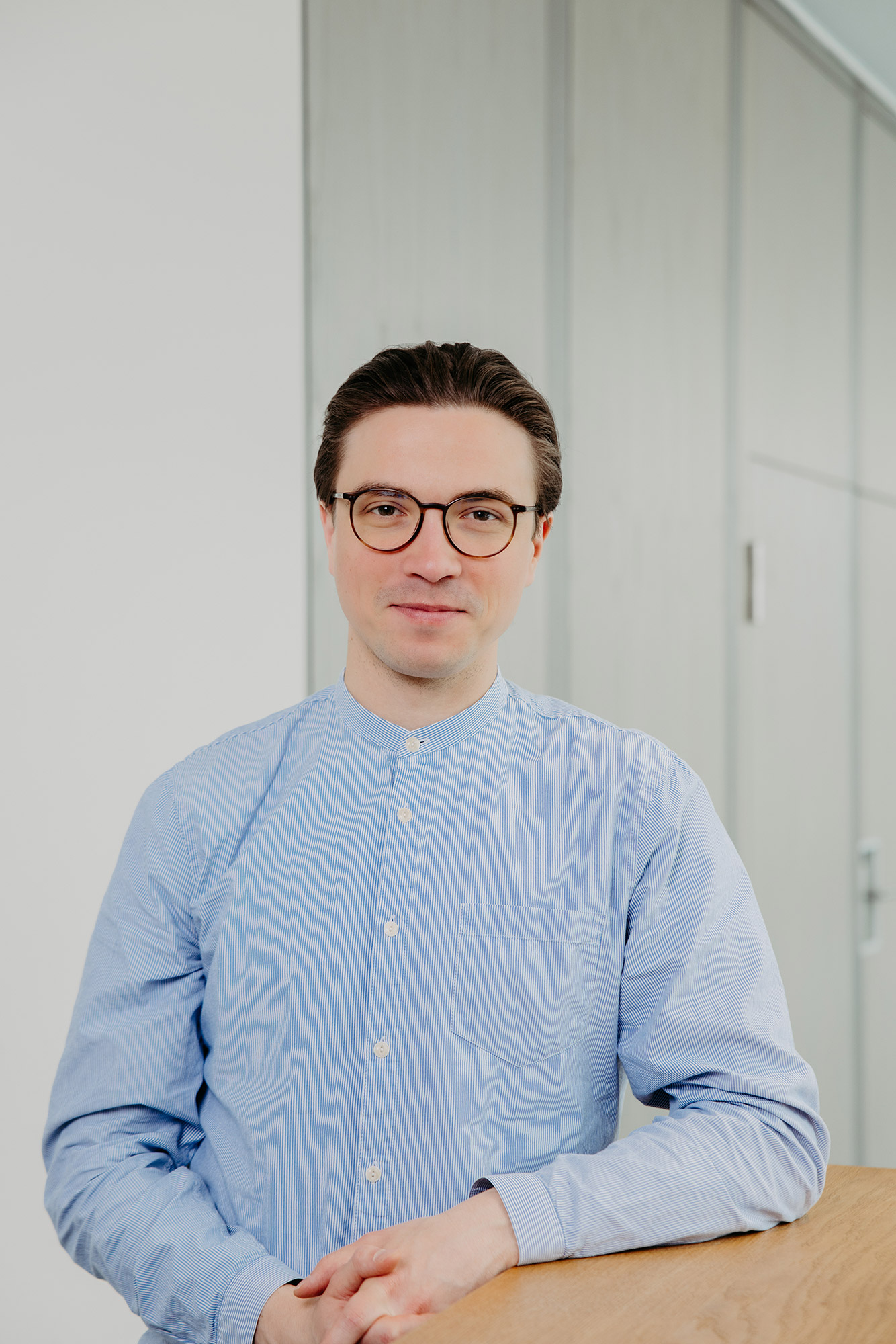 Mann mit Brille im hellblauen Hemd sitzt an einem Holztisch vor einer hellen Wand