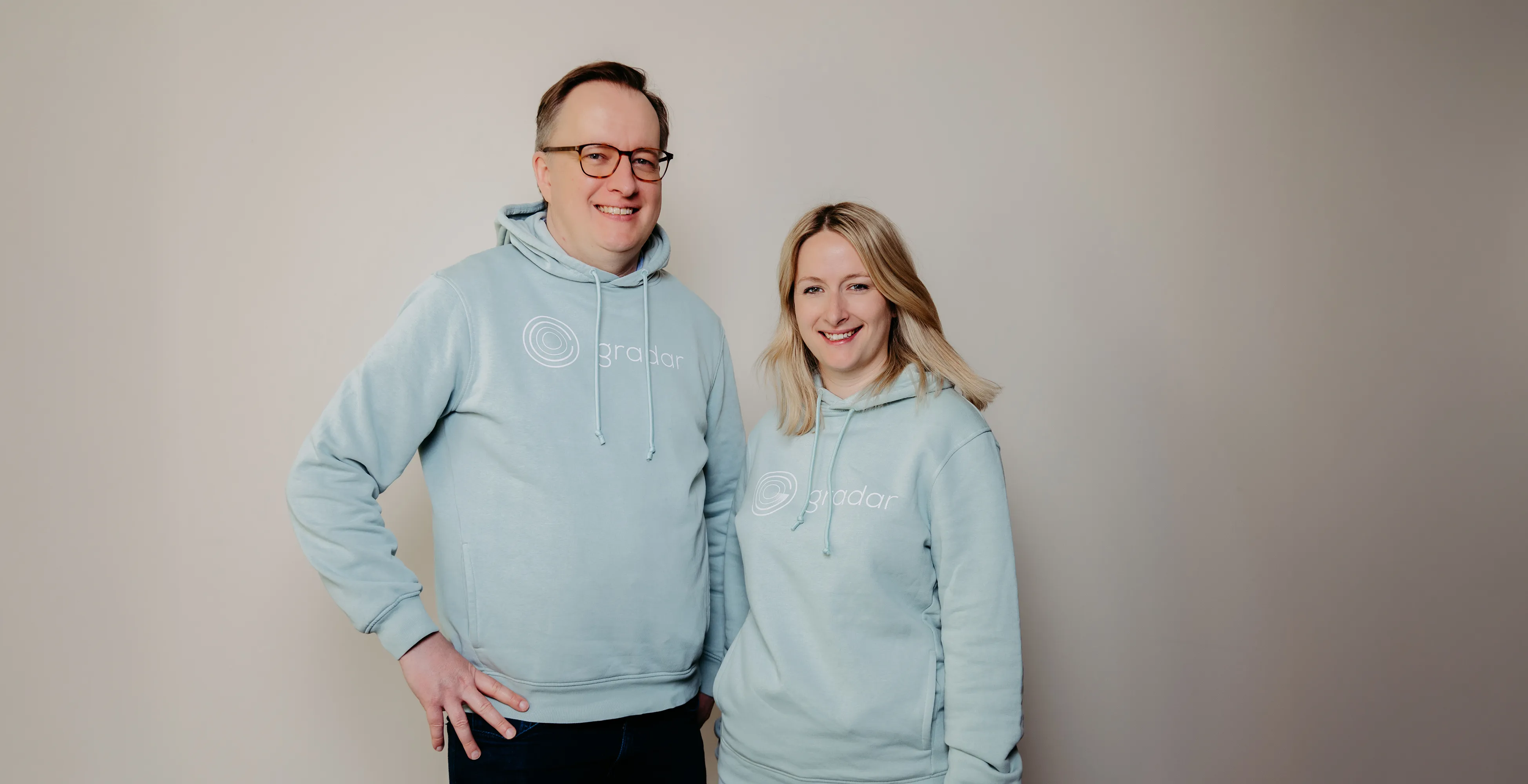 Lächelnder Mann und lächelnde Frau in hellblauen Hoodies mit gradar-Logo vor neutralem Hintergrund