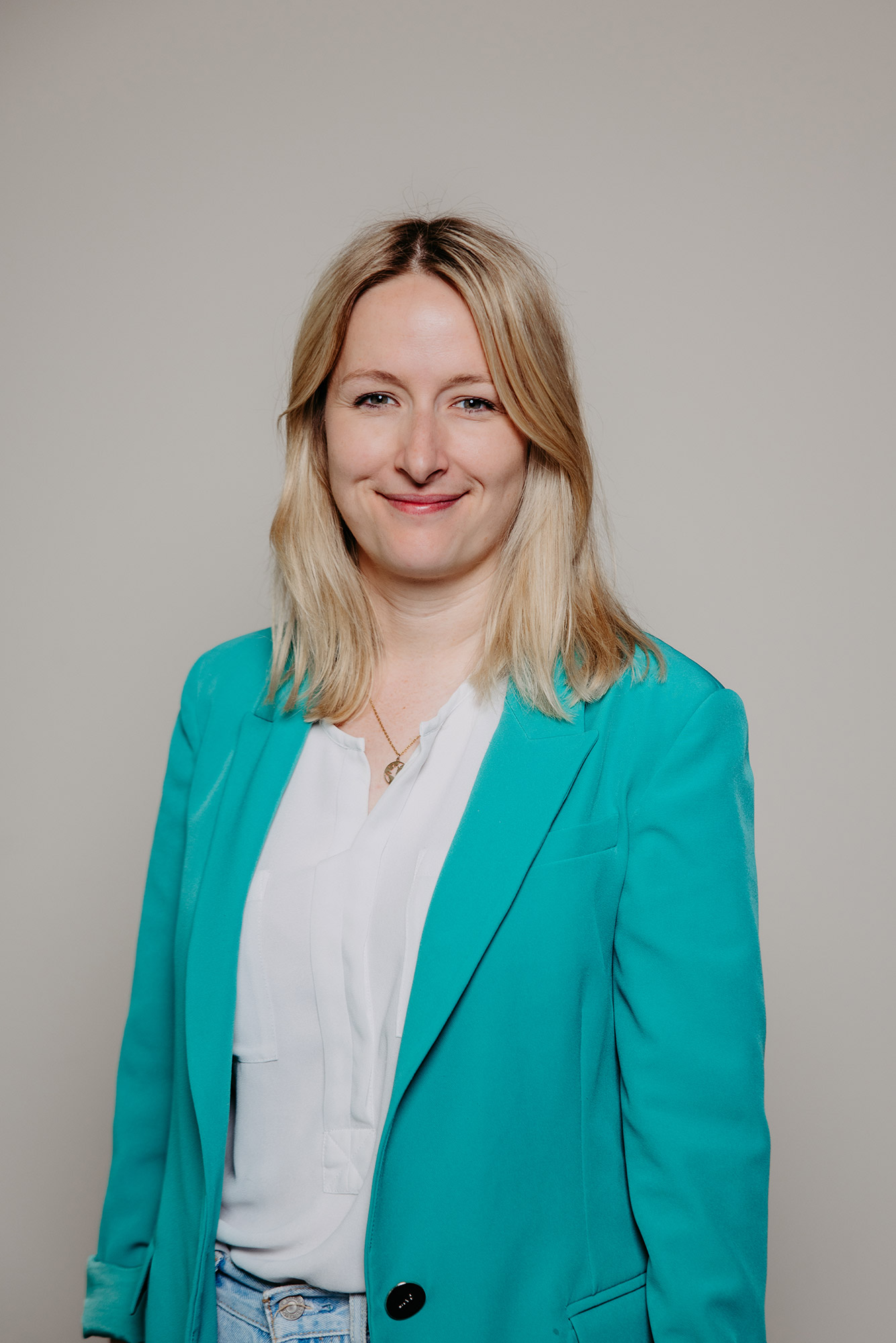 Smiling woman with blonde hair wearing a turquoise blazer and white blouse against a plain background.