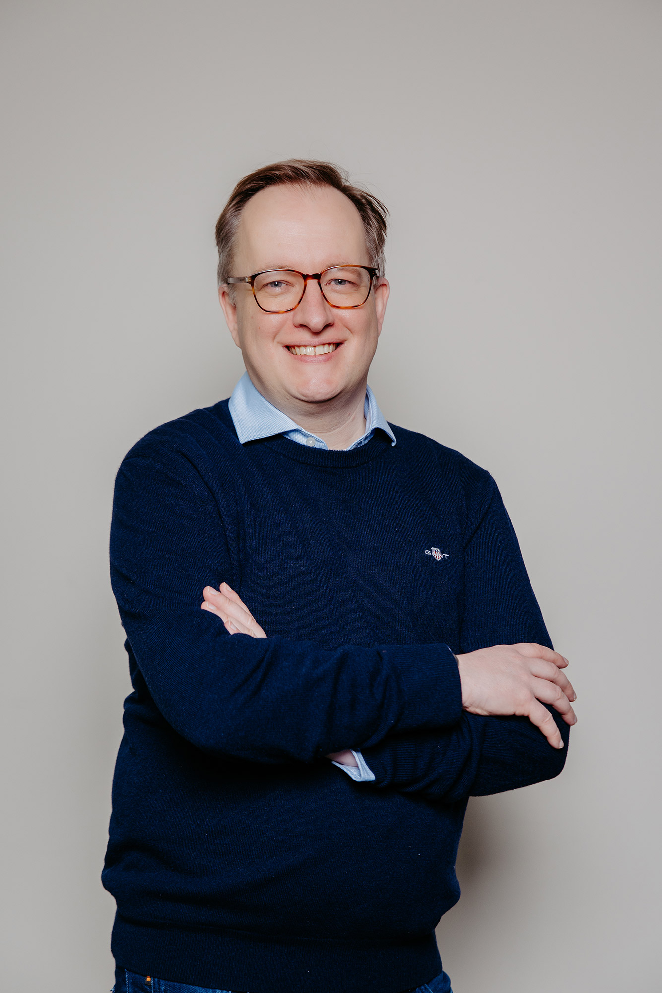 Smiling man with glasses in a navy sweater and light blue shirt posing against a plain background.