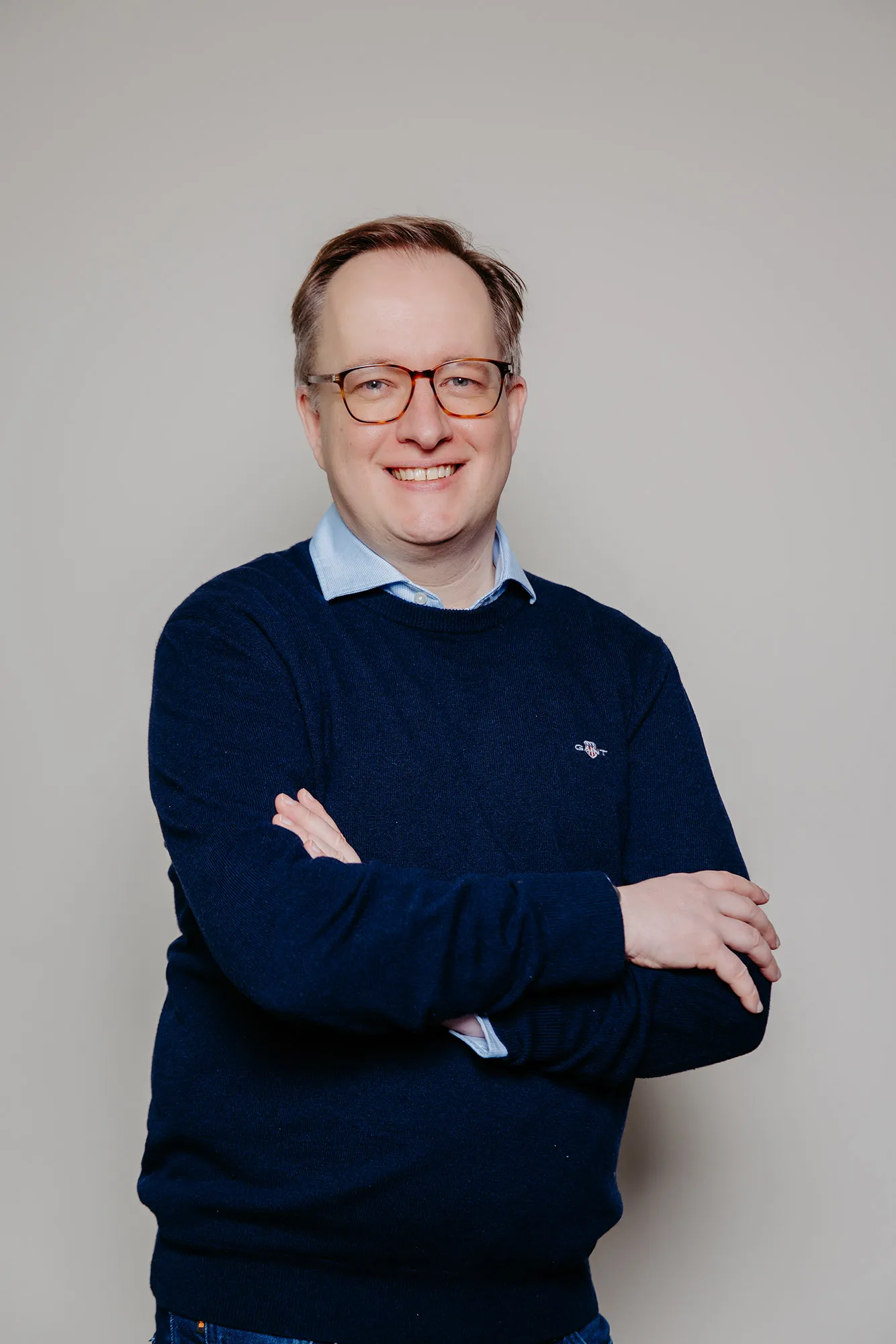 Smiling man with glasses in a navy sweater and light blue shirt posing against a plain background.