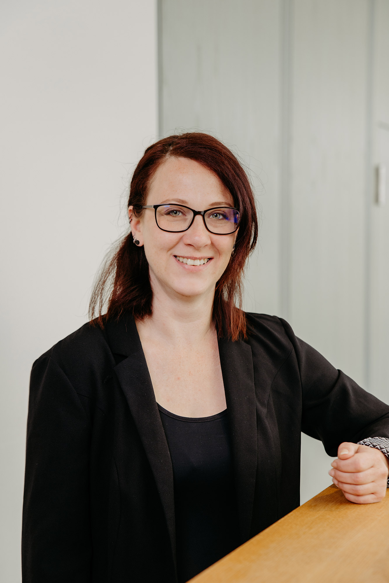 Lächelnde Frau mit Brille und roten Haaren im schwarzen Blazer, die sich auf eine Holzfläche stützt