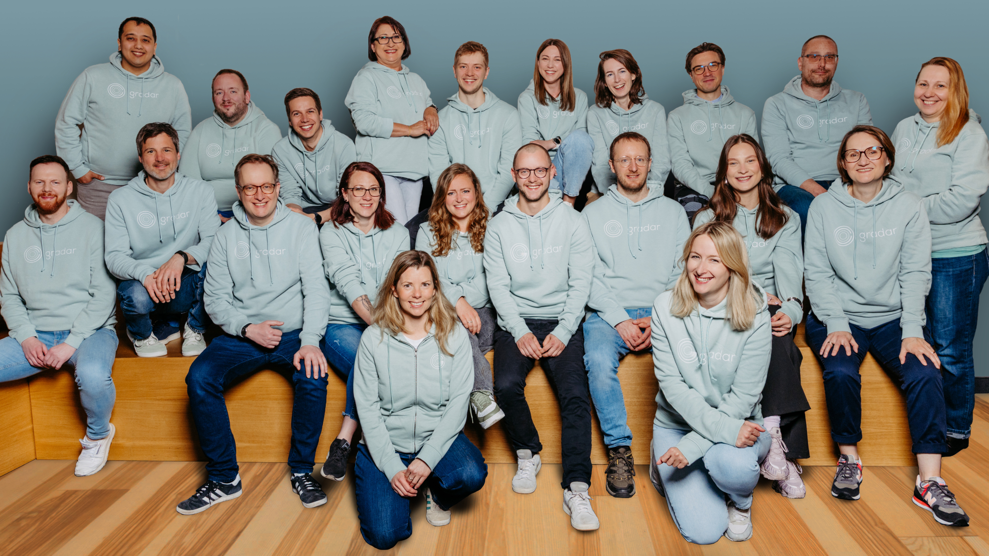 Group of 23 people wearing matching light blue hoodies sitting and kneeling on wooden stairs.