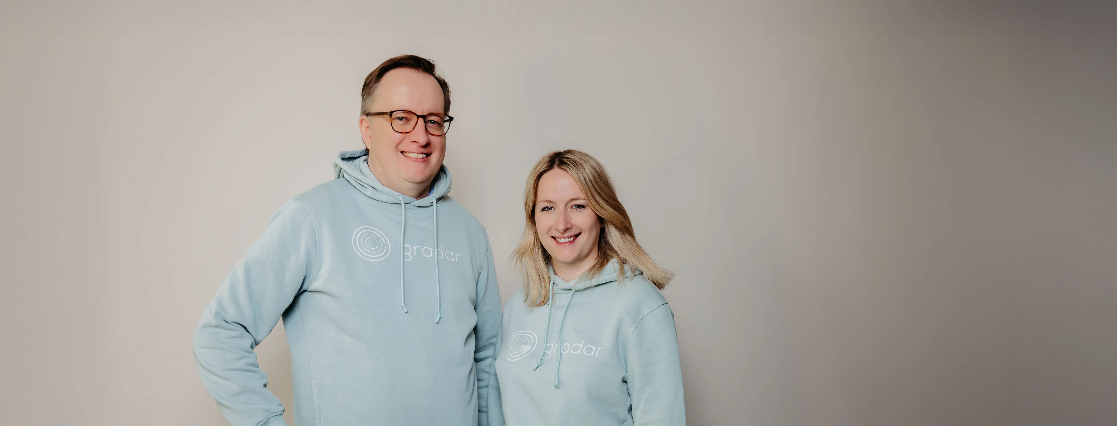 Man and woman wearing matching light blue hoodies with Gradar logo smiling against plain background.