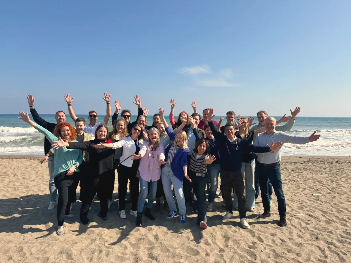 Group of smiling people standing on a sandy beach with ocean waves and blue sky behind them.