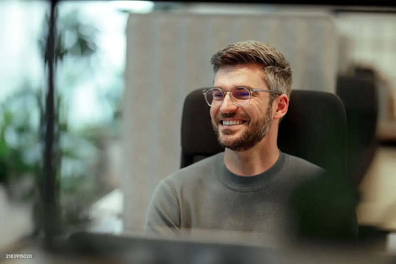 Smiling man with glasses and beard sitting in an office chair by a window with plants outside.