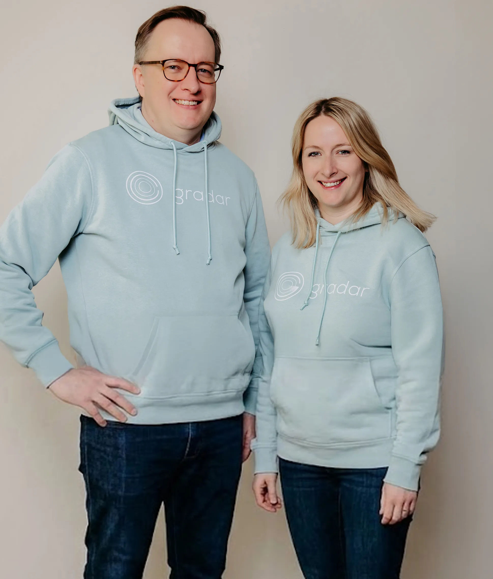 A man and a woman standing side by side against a plain background, both smiling and wearing light blue hoodies with the word 'gradar' and a circular logo printed on them and dark blue jeans.