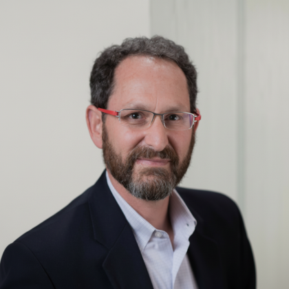 Man with short curly dark hair and beard wearing glasses with red arms, a white shirt, and a dark blazer, smiling slightly against a neutral background.