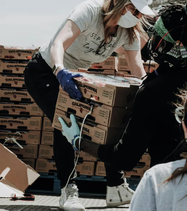 Two masked and gloved people lifting stacked boxes labeled California, surrounded by more boxes on pallets outdoors.