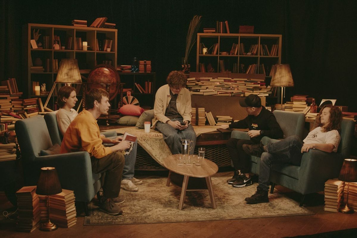 Fünf Personen sitzen in einem gemütlichen Raum mit Büchern, zwei Sesseln und einem Couchtisch mit Gläsern.