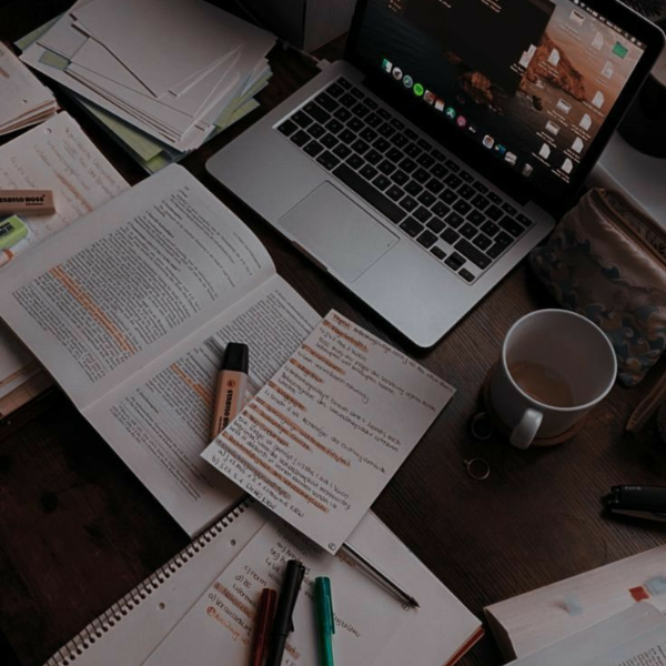 Arbeitsplatz mit offenem Laptop, mehreren Büchern, Notizblöcken, Stiften und Tasse Kaffee auf einem Holzschreibtisch.