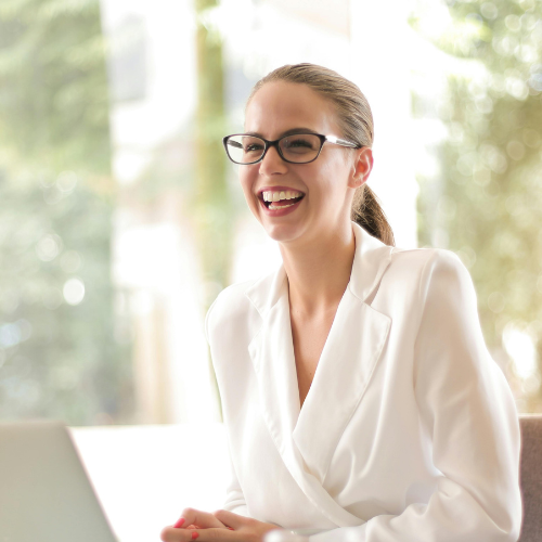 Lachende Frau mit Brille und weißem Blazer sitzt vor einem Laptop in einem hellen Raum.
