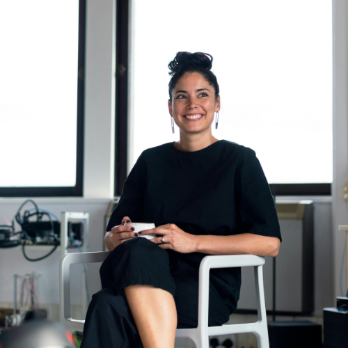 Frau mit schwarzer Kleidung und hochgestecktem Haar lächelt und hält eine Tasse, während sie auf einem Stuhl sitzt.