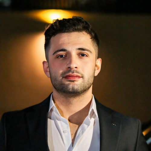 Porträt eines jungen Mannes mit dunklem Haar und leichtem Bart in weißem Hemd und dunklem Sakko vor unscharfem Hintergrund mit warmem Licht.