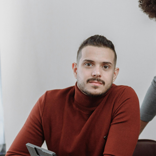 Junger Mann mit kurzem dunklem Haar und Bart trägt roten Rollkragenpullover und schaut in die Kamera.