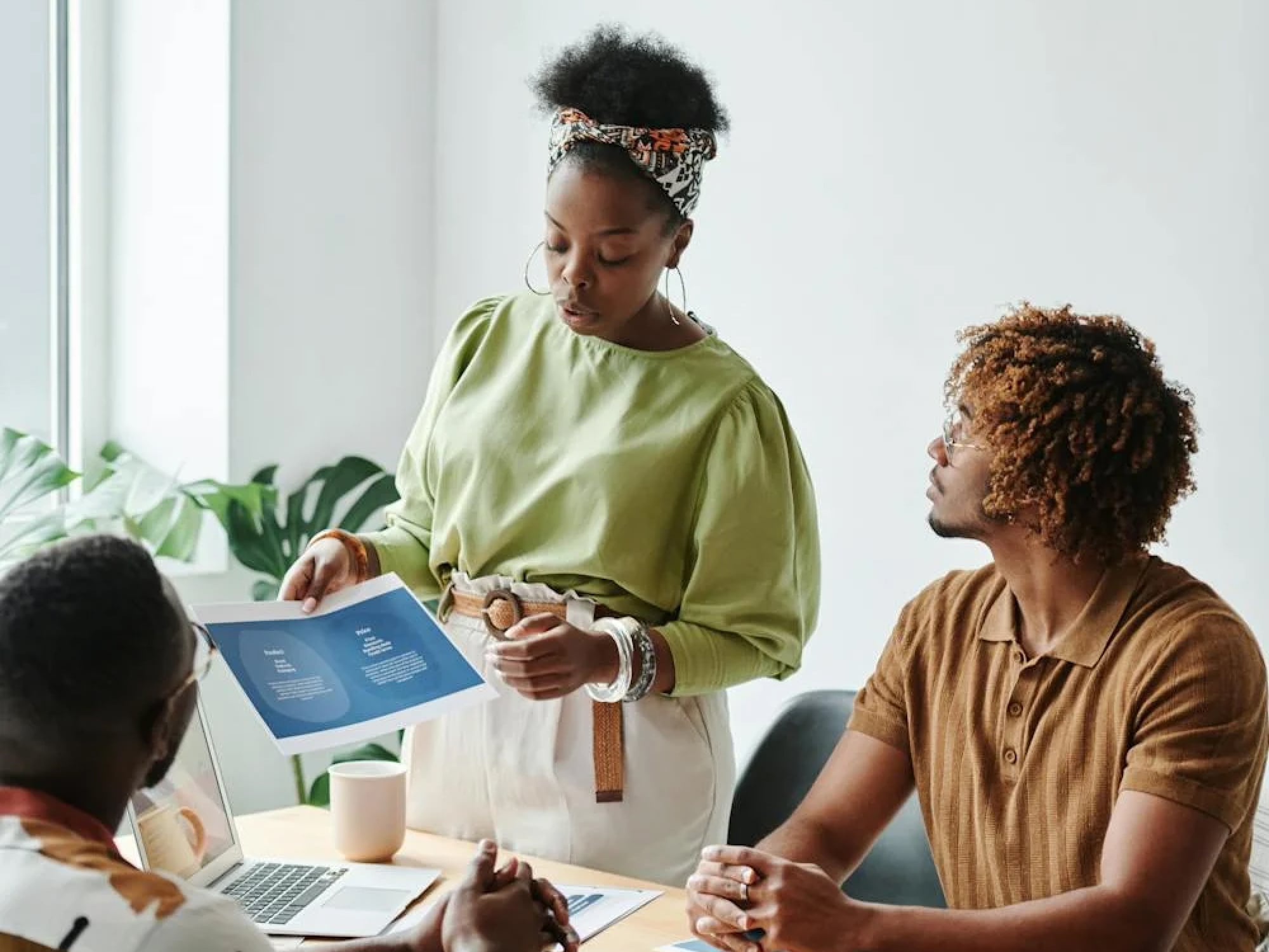 Une femme présente un document à deux hommes assis autour d'une table de réunion avec un ordinateur portable et une tasse.