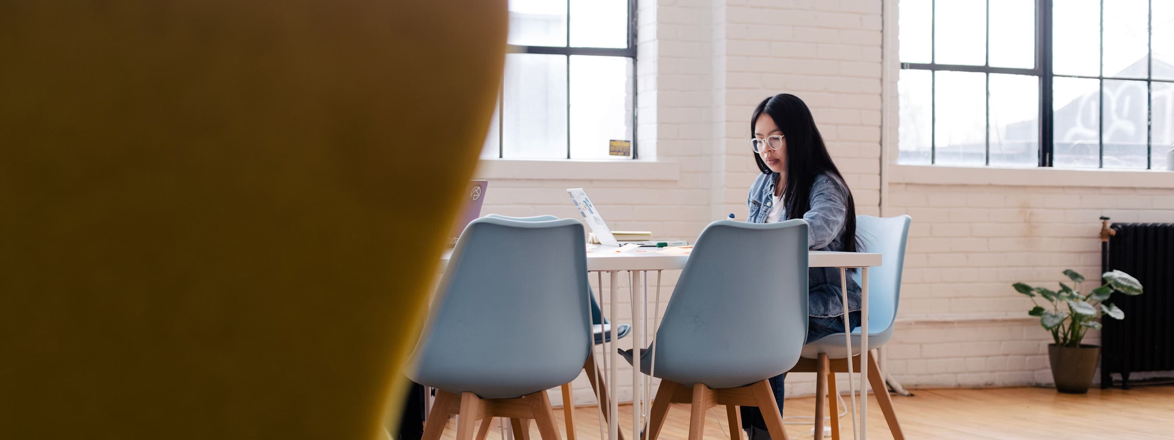 Une femme aux cheveux longs et lunettes travaille à une table blanche dans un bureau lumineux avec des chaises bleues et une plante verte.