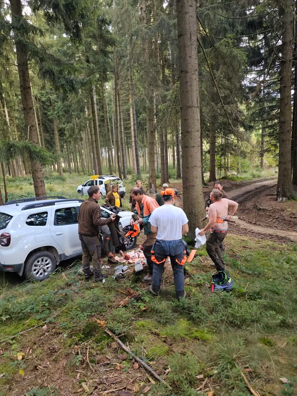 Eine Gruppe von Männern steht in einem Wald um eine weiße SUV herum, einige tragen Arbeitskleidung, einer ist oberkörperfrei.
