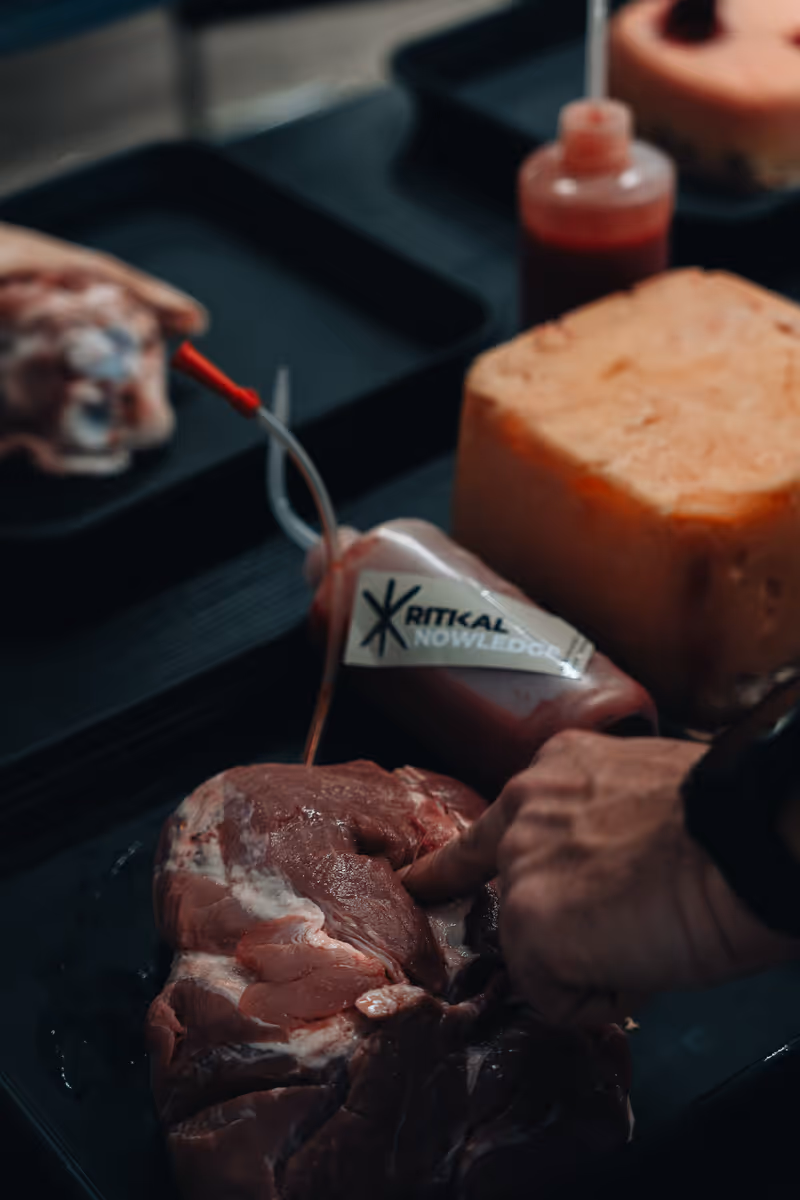 Hand berührt rohes Fleisch neben einem Blutbeutel mit Aufschrift auf schwarzem Tablett.