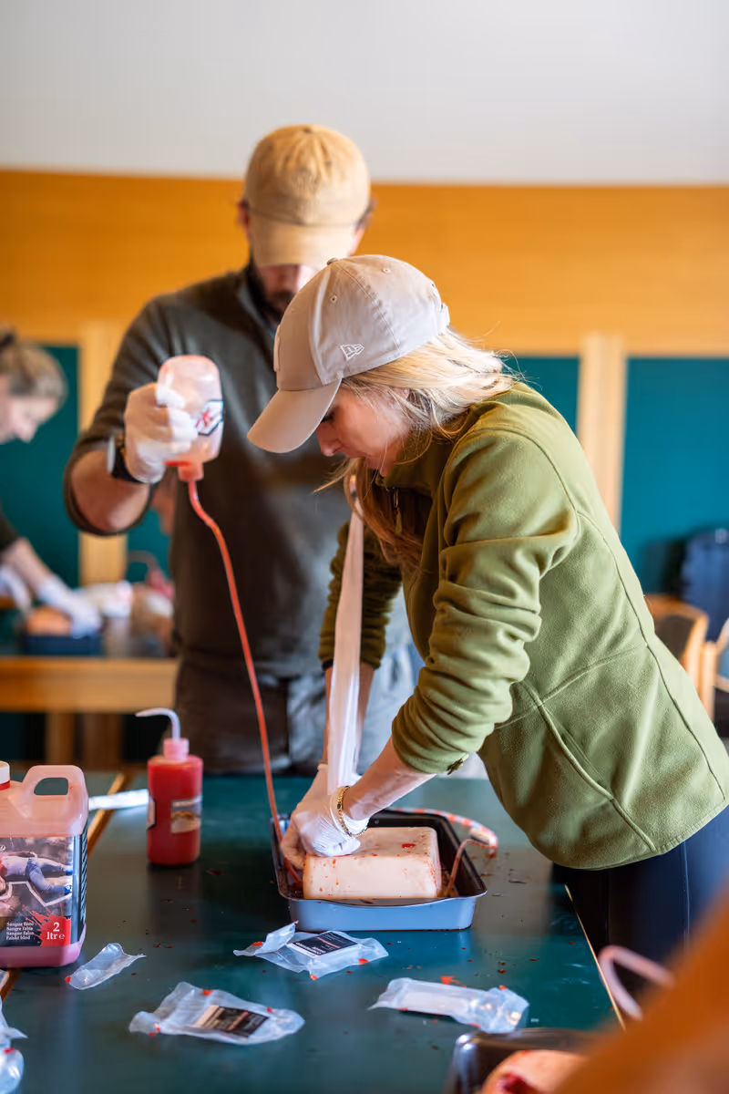 Frau mit Basecap und grüner Jacke arbeitet mit Material in einer Schale auf einem Tisch, während ein Mann im Hintergrund eine rote Flüssigkeit aus einer Flasche gießt.
