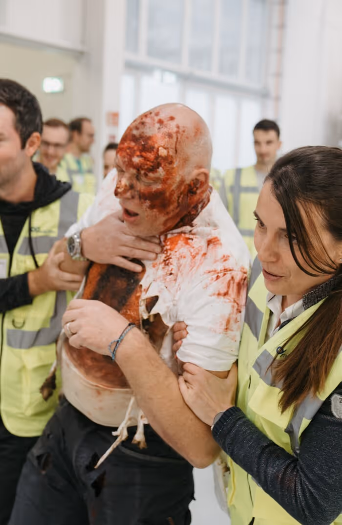 Mehrere Sanitäter in neongelben Westen stützen einen Mann mit blutverschmierter Kleidung und Gesichtsverletzungen bei Rettungsübung.