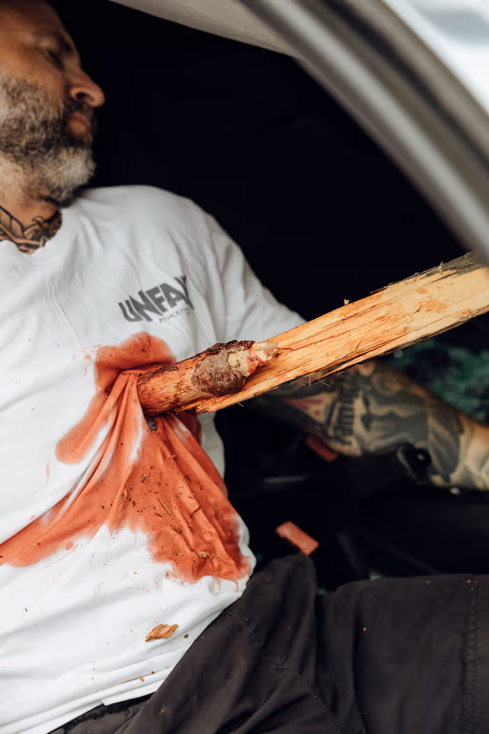 Ein Mann mit Bart und Tätowierungen sitzt, und ein großes Holzstück steckt durch sein weißes Hemd, das blutbefleckt ist.