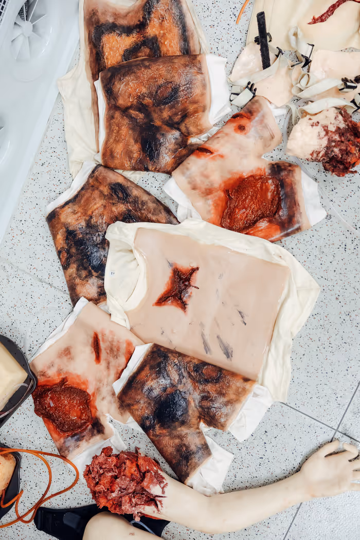 Mehrere realistisch aussehende Wund-Simulationsmasken und eine künstliche verletzte Hand liegen auf einem hellen gefliesten Boden.