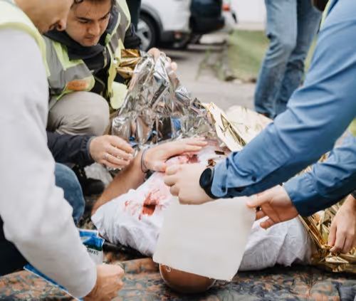 Mehrere Personen, die einer verwundeten Person mit einer Rettungsdecke und Erste-Hilfe-Maßnahmen im Freien helfen.