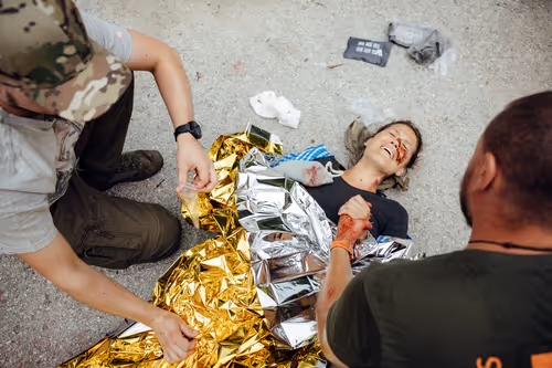 Zwei Personen helfen einer verletzten Frau, die auf dem Boden liegt und in eine Rettungsdecke gewickelt wird.