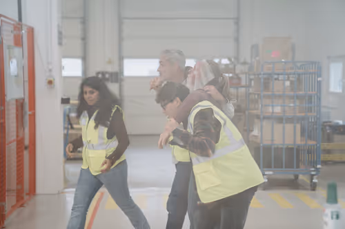 Drei Personen in Warnwesten helfen einem verletzten Kollegen in einem Lagerhaus.