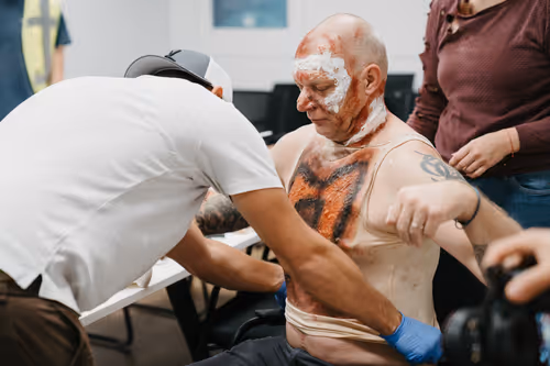 Ein Mann mit Makeup und Kunstblut auf Kopf und Oberkörper wird von zwei Personen für eine Simulation medizinisch behandelt.